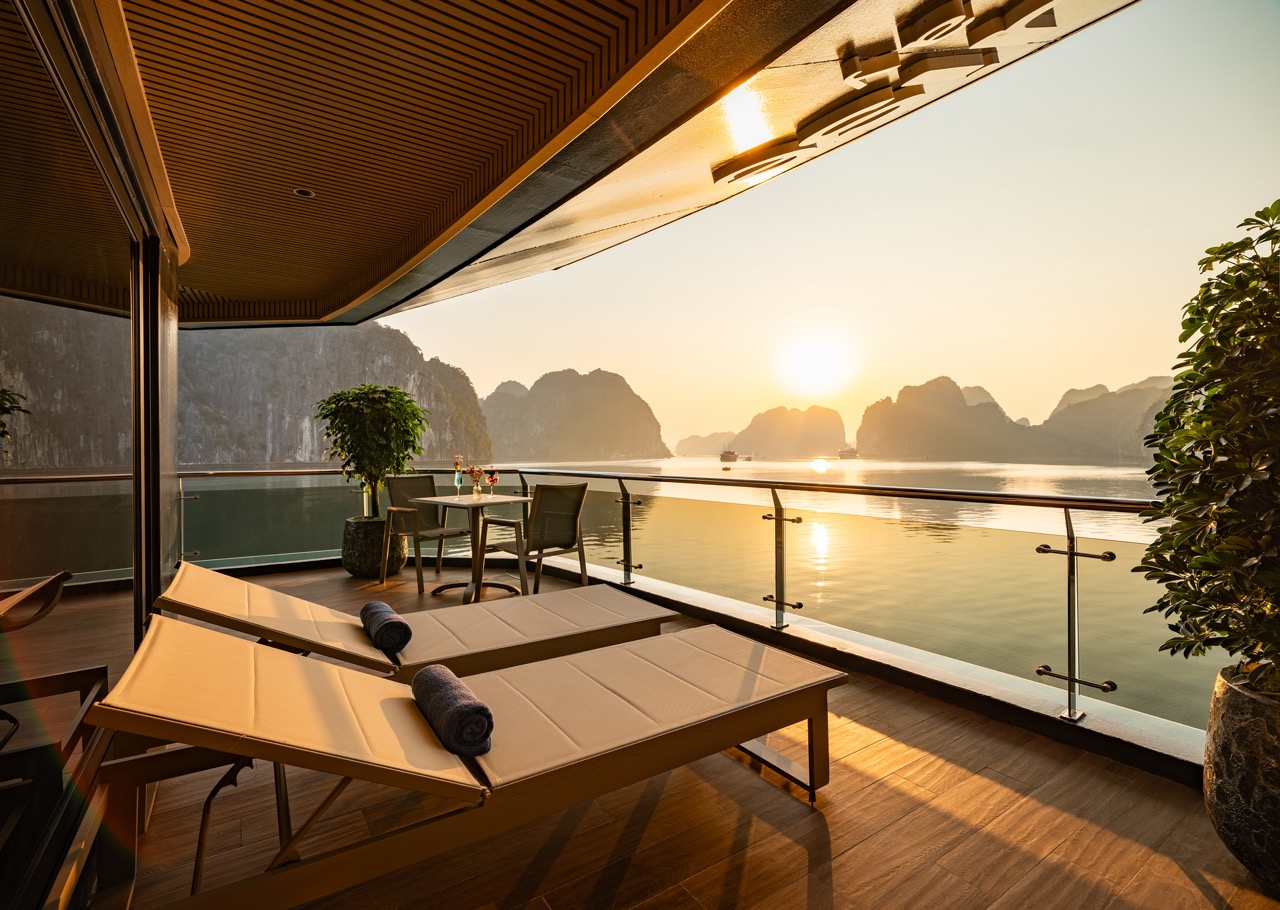 Sunset view from a luxury yacht balcony with two beige lounge chairs, a small table, and glass railing overlooking calm water and distant limestone islands.