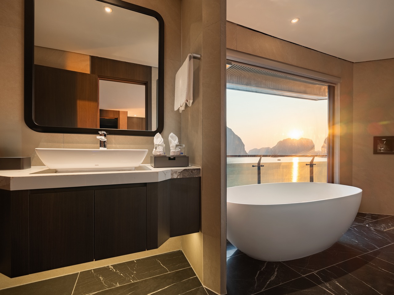 Contemporary bathroom with a white freestanding tub by a large window overlooking a sunset over water and mountains, and a dark wood vanity with a vessel sink.