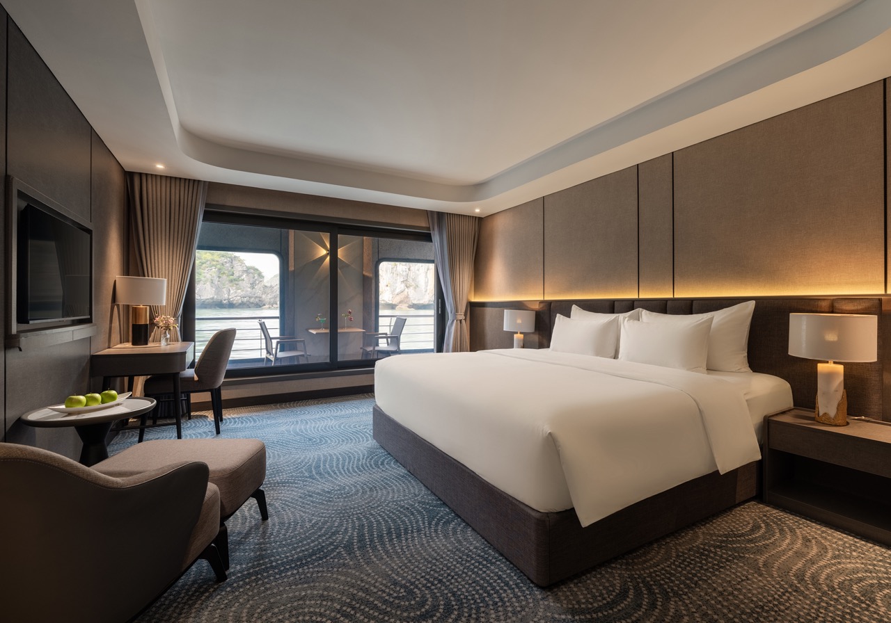 King-size bed in a modern hotel room with ambient lighting, nightstands, and a balcony with outdoor seating overlooking water and rocks.