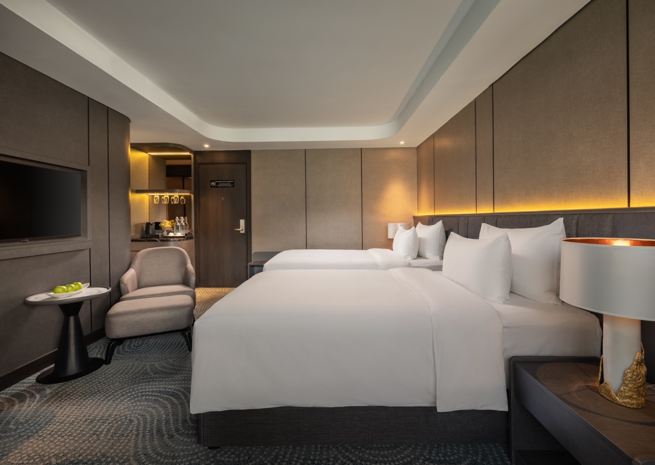 Hotel room with two large white beds, a gray headboard, warm ambient lighting, and a seating area nearby.