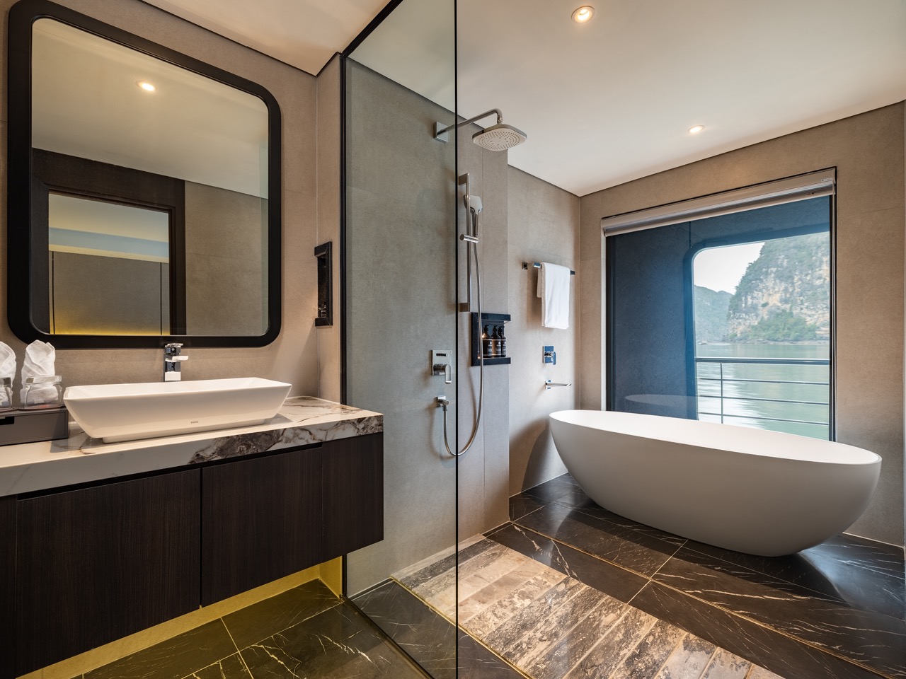 Contemporary bathroom with a freestanding oval tub by a large window overlooking water and cliffs; glass shower and dark vanity.