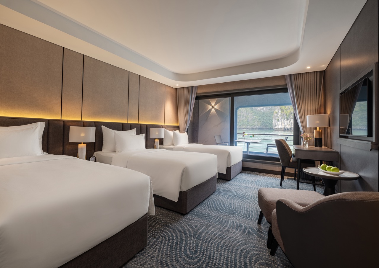 Hotel room with three neatly made white beds, a beige padded headboard, warm bedside lamps, and a large window overlooking water and cliffs.