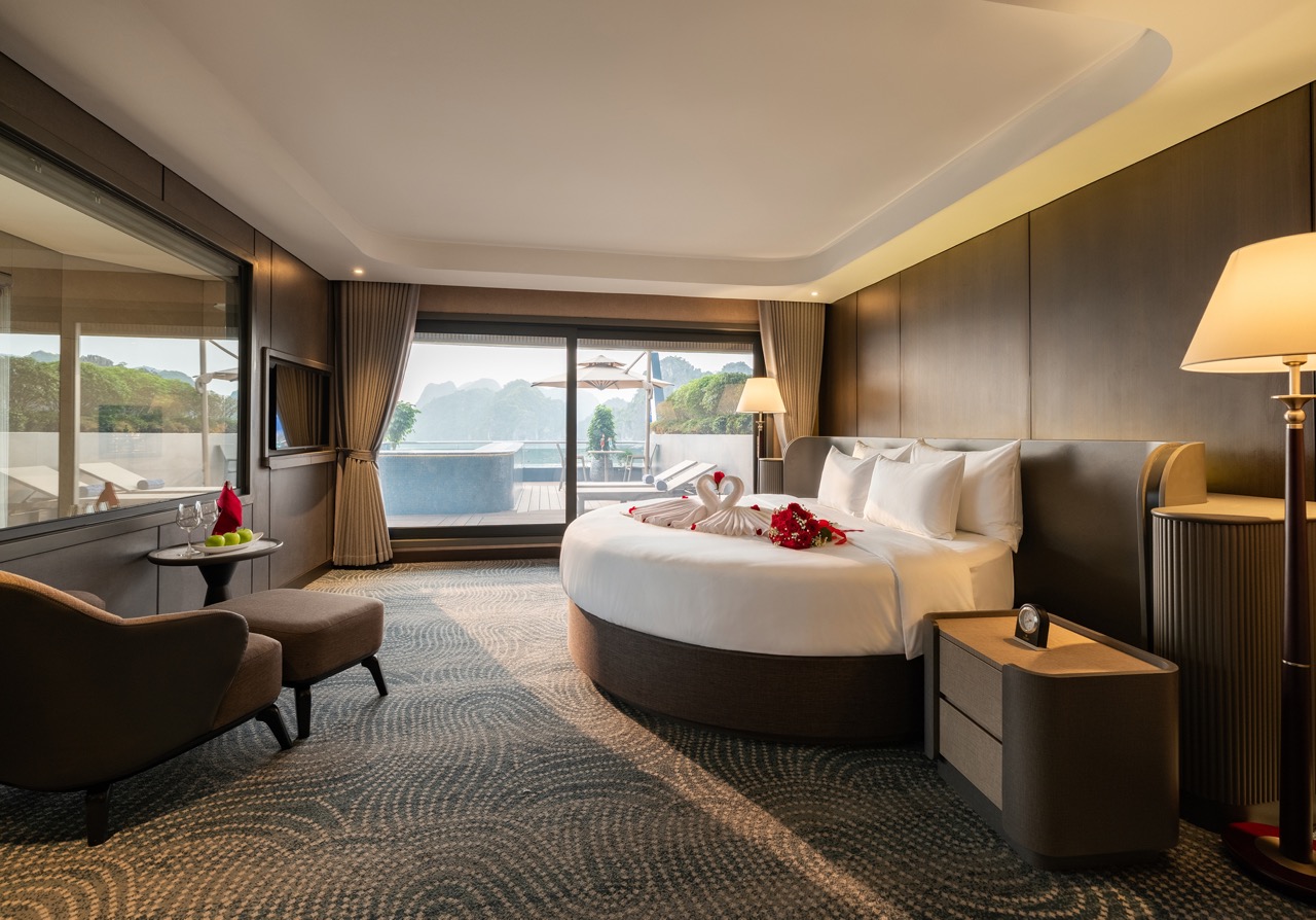 Luxury hotel room with a round king bed, towel swans, and rose petals, opening to a terrace with mountain view.