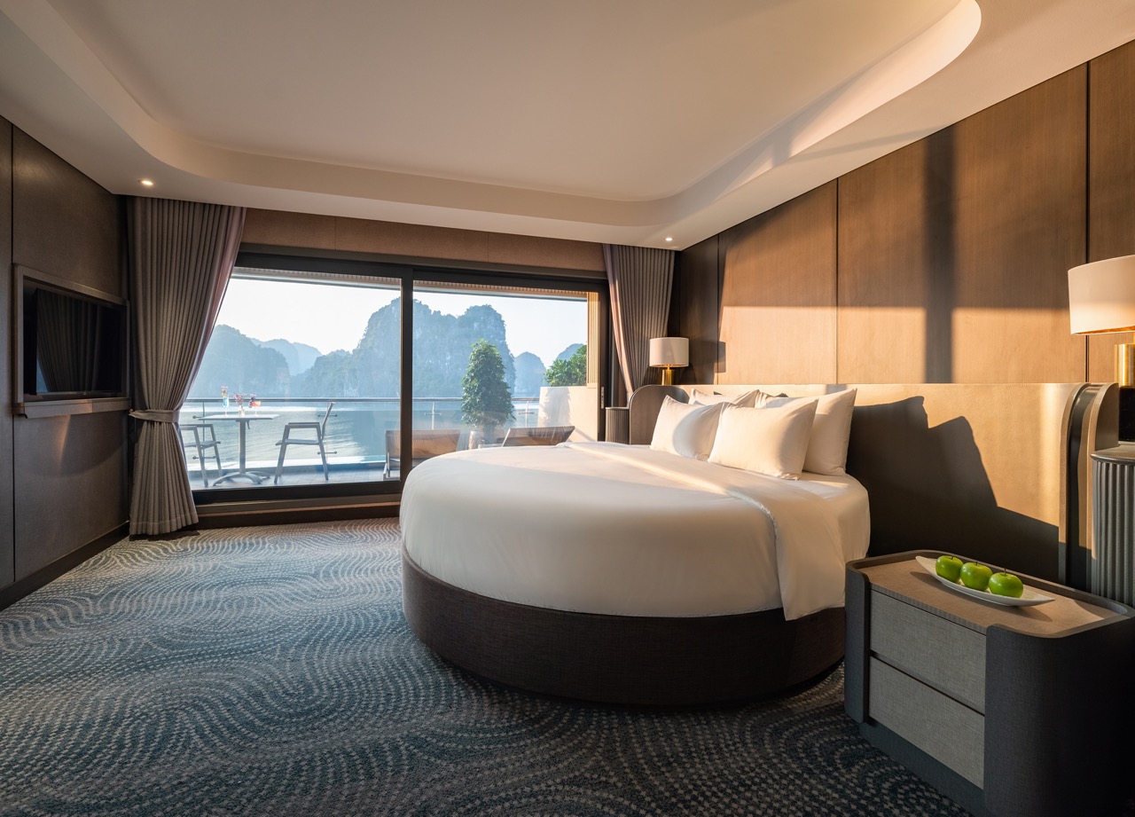 Luxurious hotel room featuring a round bed, bedside tables and lamps, with a balcony and sea-cliff view through a large window/doorway.