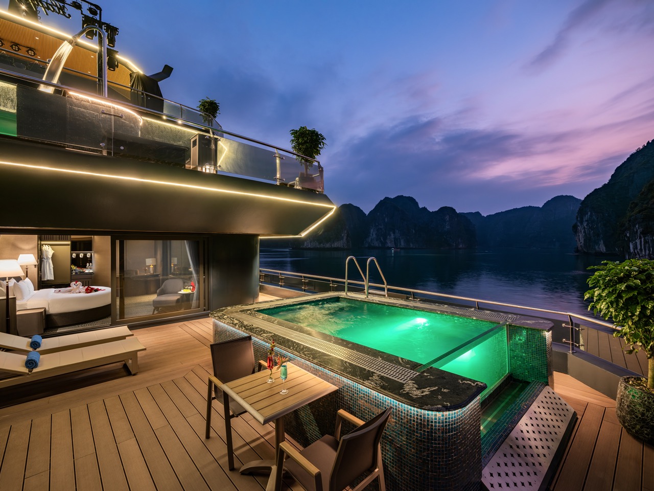 Yacht deck at sunset with a glowing pool, loungers, and a glass-walled bedroom visible inside, across calm blue water and limestone cliffs.