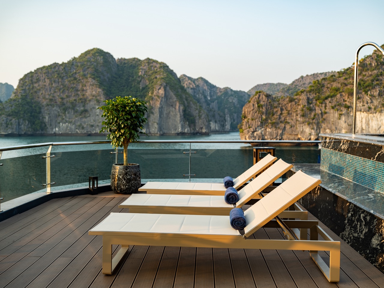 Outdoor pool deck with three white lounge chairs and blue towels, facing a bay with rocky cliffs and calm water behind a glass railing.