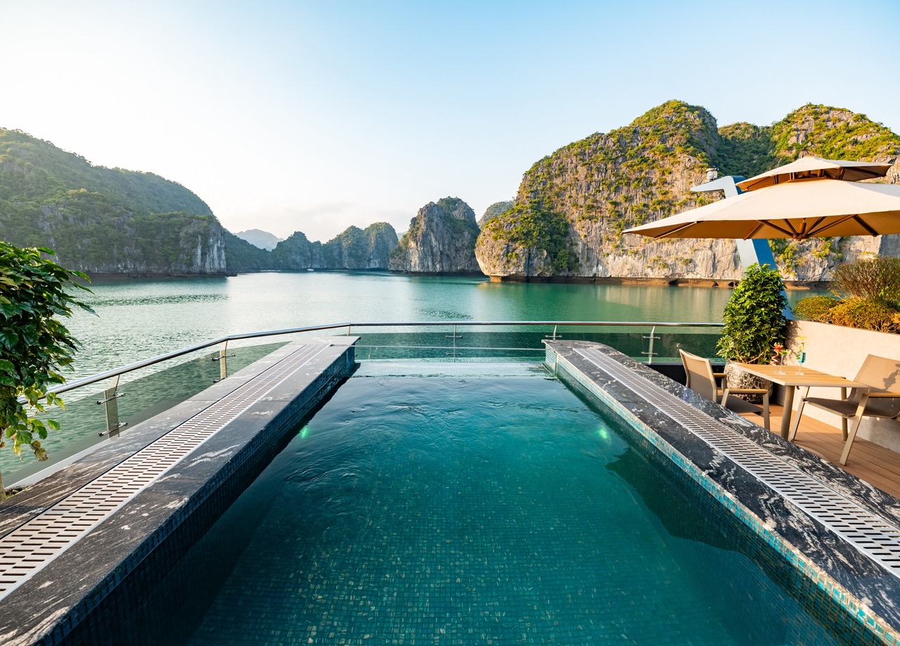 Infinity pool on a terrace overlooking a turquoise bay with limestone cliffs and distant hills; seating and umbrellas on the right side.