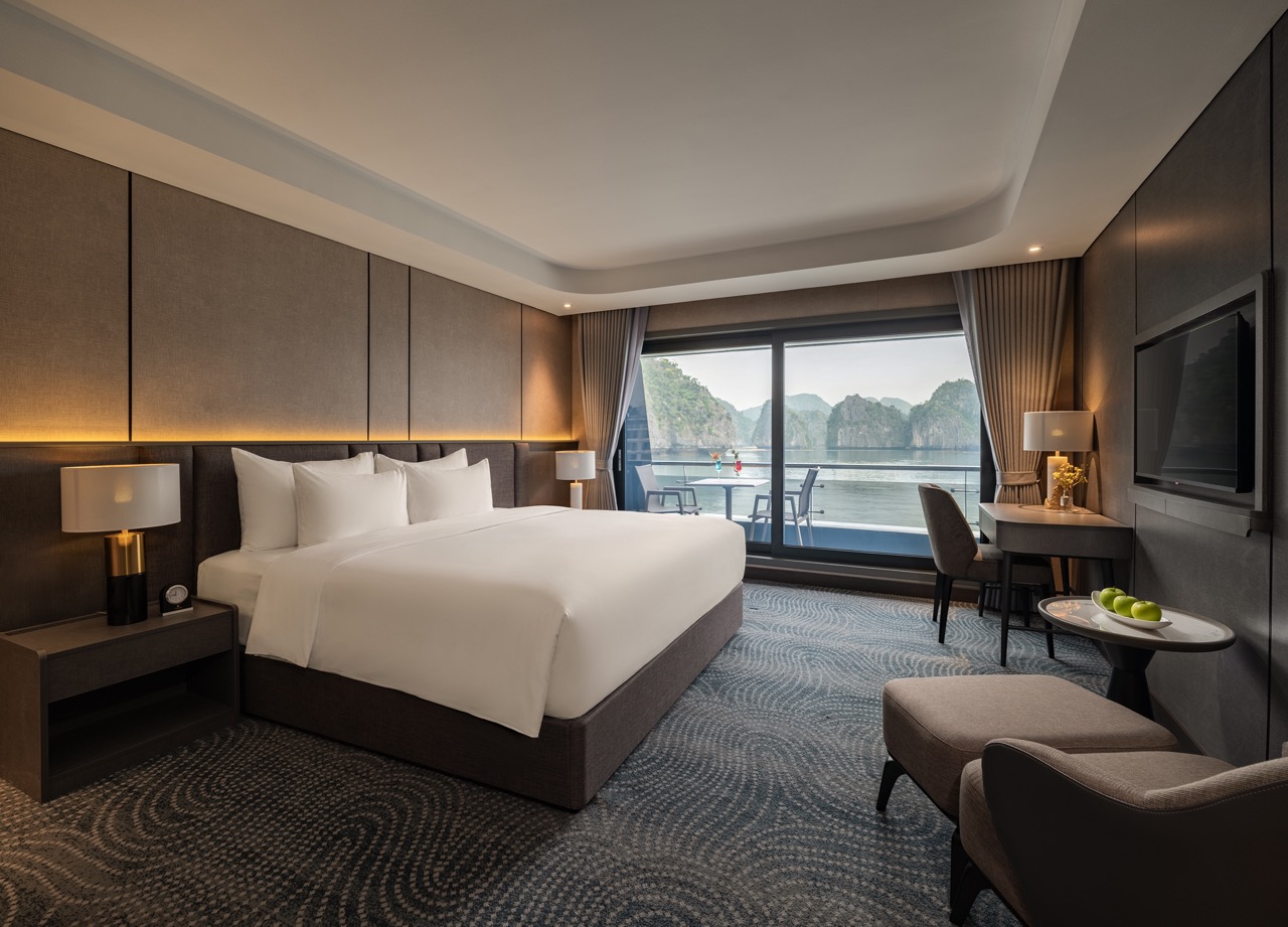 Modern hotel room with a king bed, plush white bedding, and twin nightstands flanking the headboard; large glass doors open to a balcony with water and rocky islands outside.