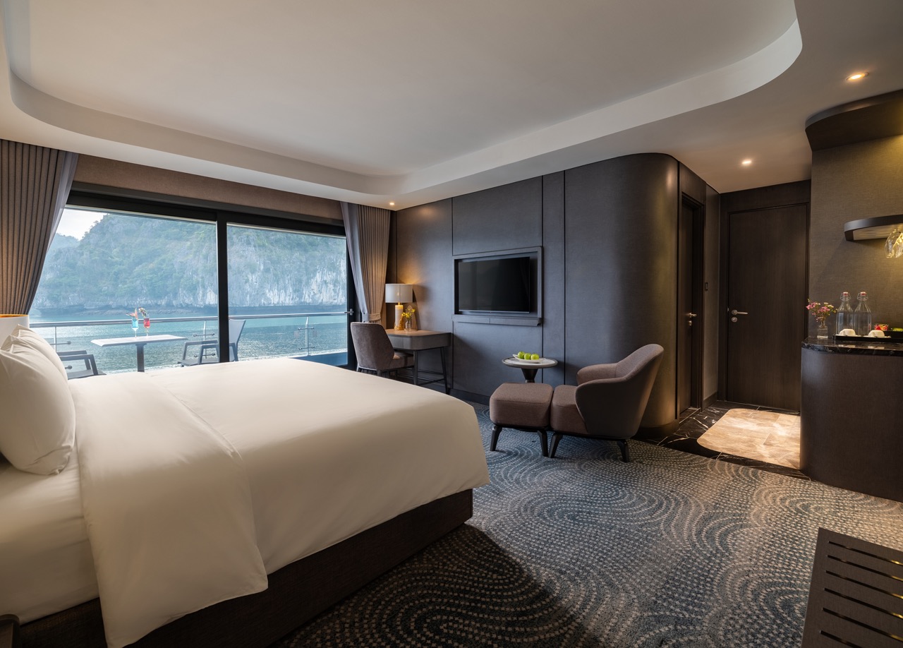 Luxurious hotel suite with a king bed facing a glass balcony door overlooking the ocean and rocky shoreline outside.