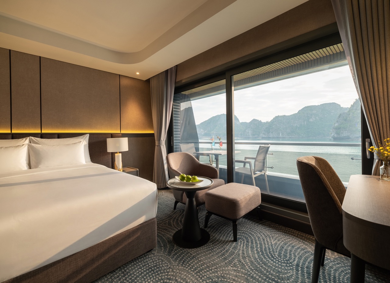 Hotel room with a king bed, soft lighting, and a balcony overlooking a calm bay with rocky islands outside.