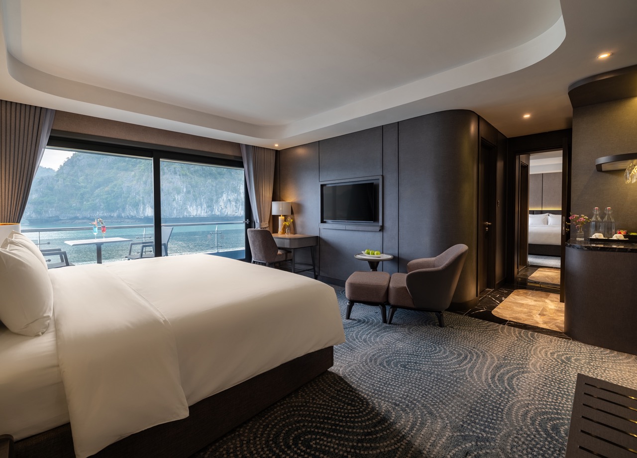 Luxurious hotel suite with a king bed, sitting area, and balcony overlooking the ocean and cliffs outside the glass doors.