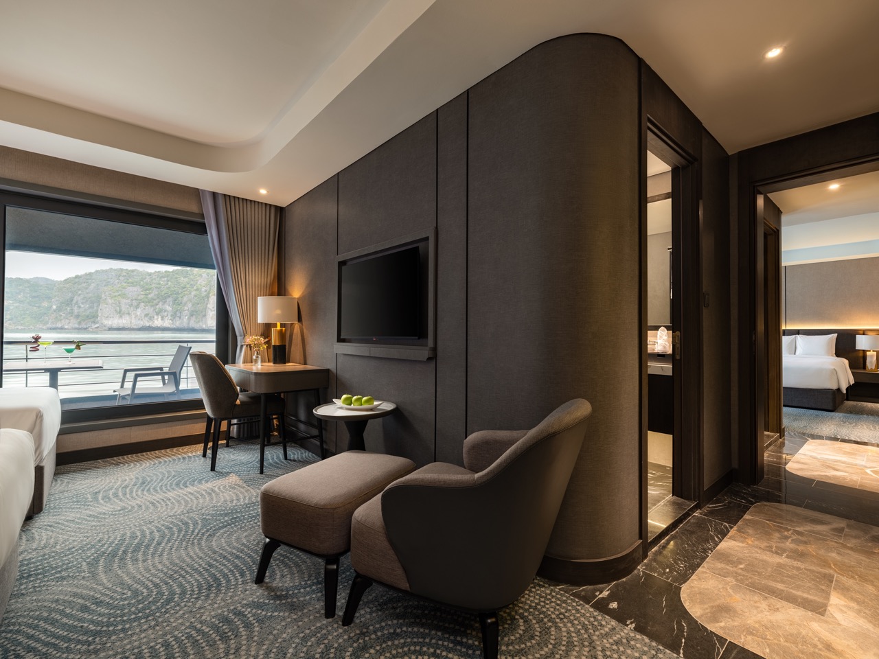 Hotel suite living area with a dark accent wall, TV, and a balcony overlooking the sea; plush chair, ottoman, and a desk with a lamp nearby.