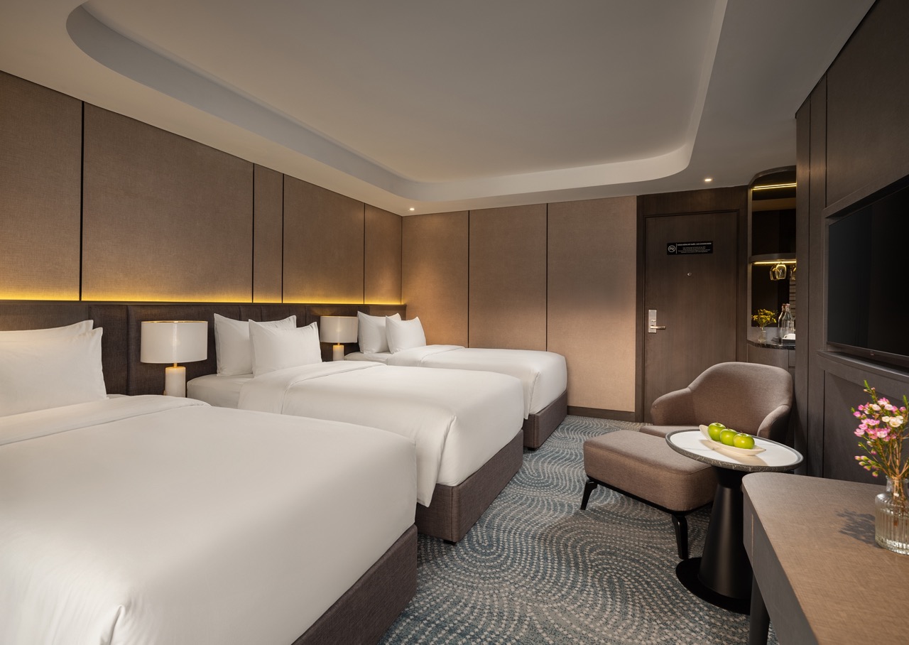 Hotel room with three white-duvet beds, beige padded headboard wall, warm bedside lamps, and a seating area with a table and fruit bowl on it