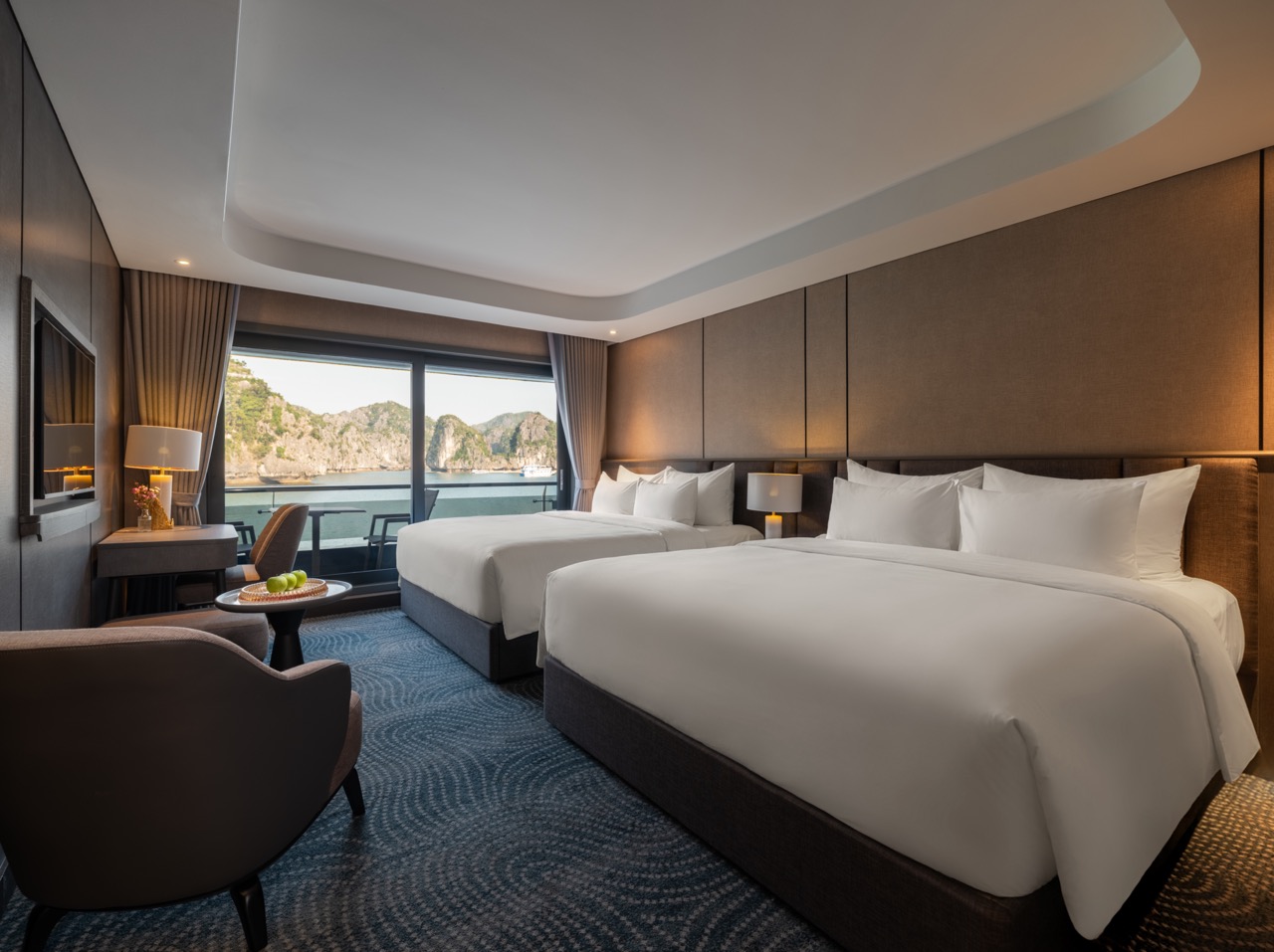 Hotel room with two large beds, seating area, and a balcony view of sea and rocky cliffs outside the window.