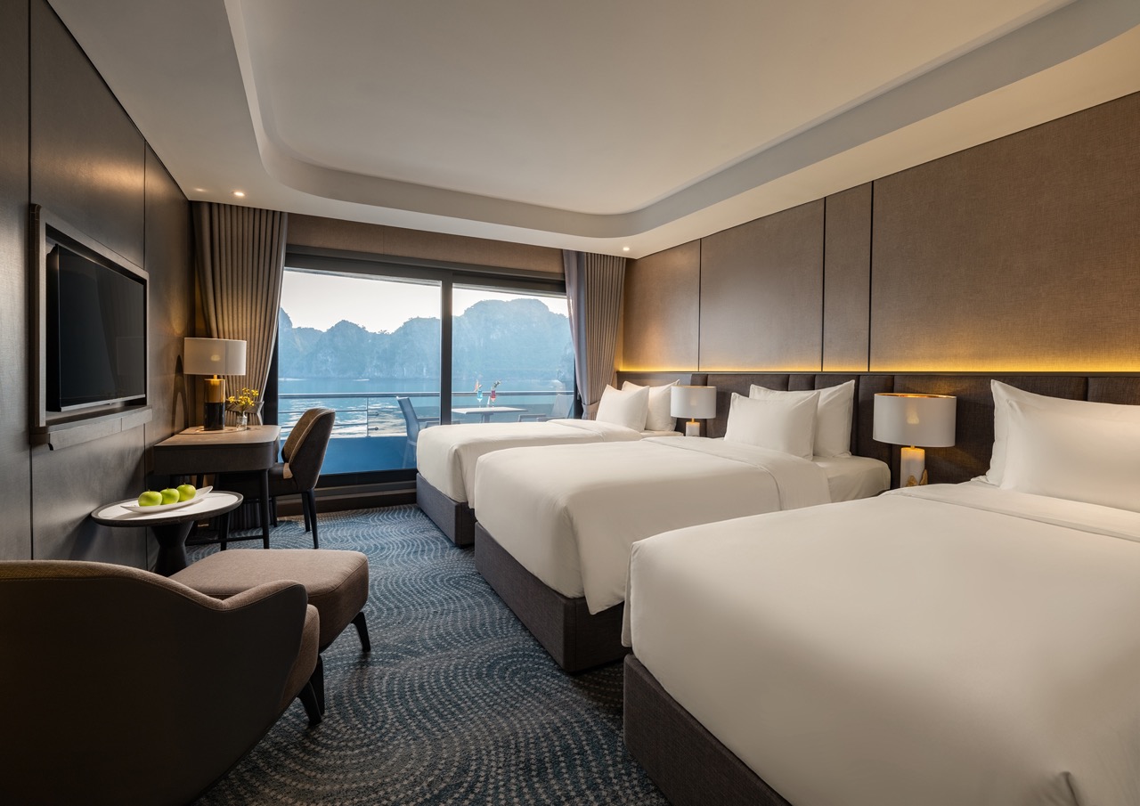 Modern hotel room with three neatly made beds, a seating area, and a balcony overlooking a rocky coastline and water beyond