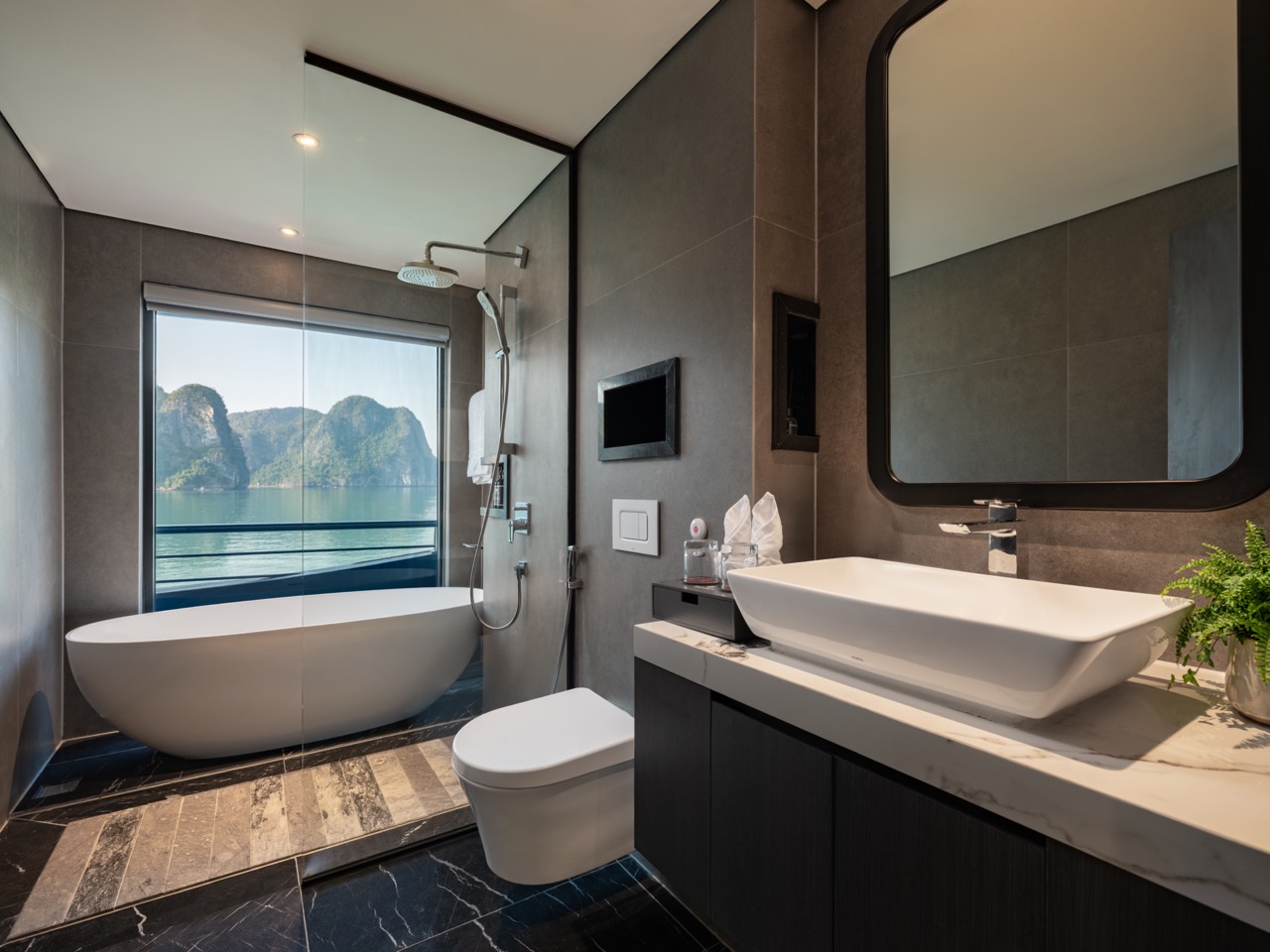 Modern bathroom with a white freestanding tub by a large window showing water and green hills, dark wall tiles, and a glass shower area.