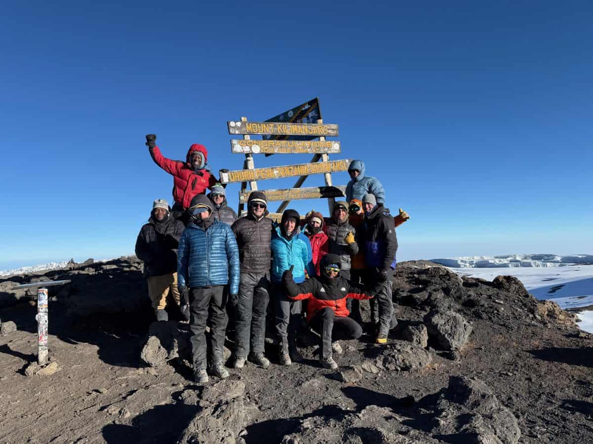 A group of climbers celebrating at a summit with a wooden sign, amidst a clear blue sky and snowy landscape.
