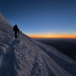 Silhouetted figures ascending a snow-covered slope at dawn, with a vibrant orange and blue sky reflecting the early morning light.