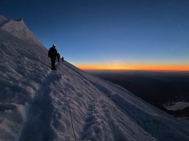 Silhouetted figures ascending a snow-covered slope at dawn, with a vibrant orange and blue sky reflecting the early morning light.