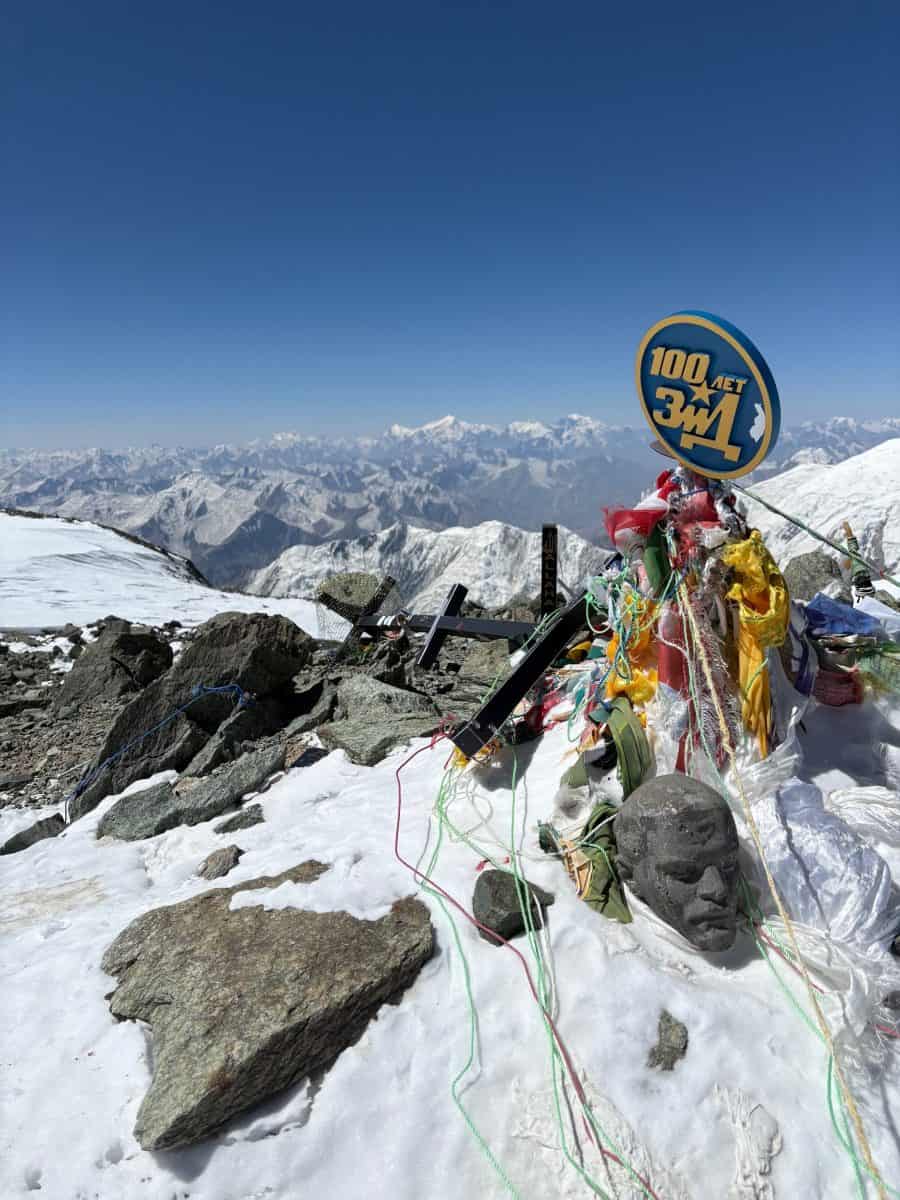 A snowy mountain peak adorned with colorful prayer flags, a stone sculpture, and a circular sign against a backdrop of distant mountains.