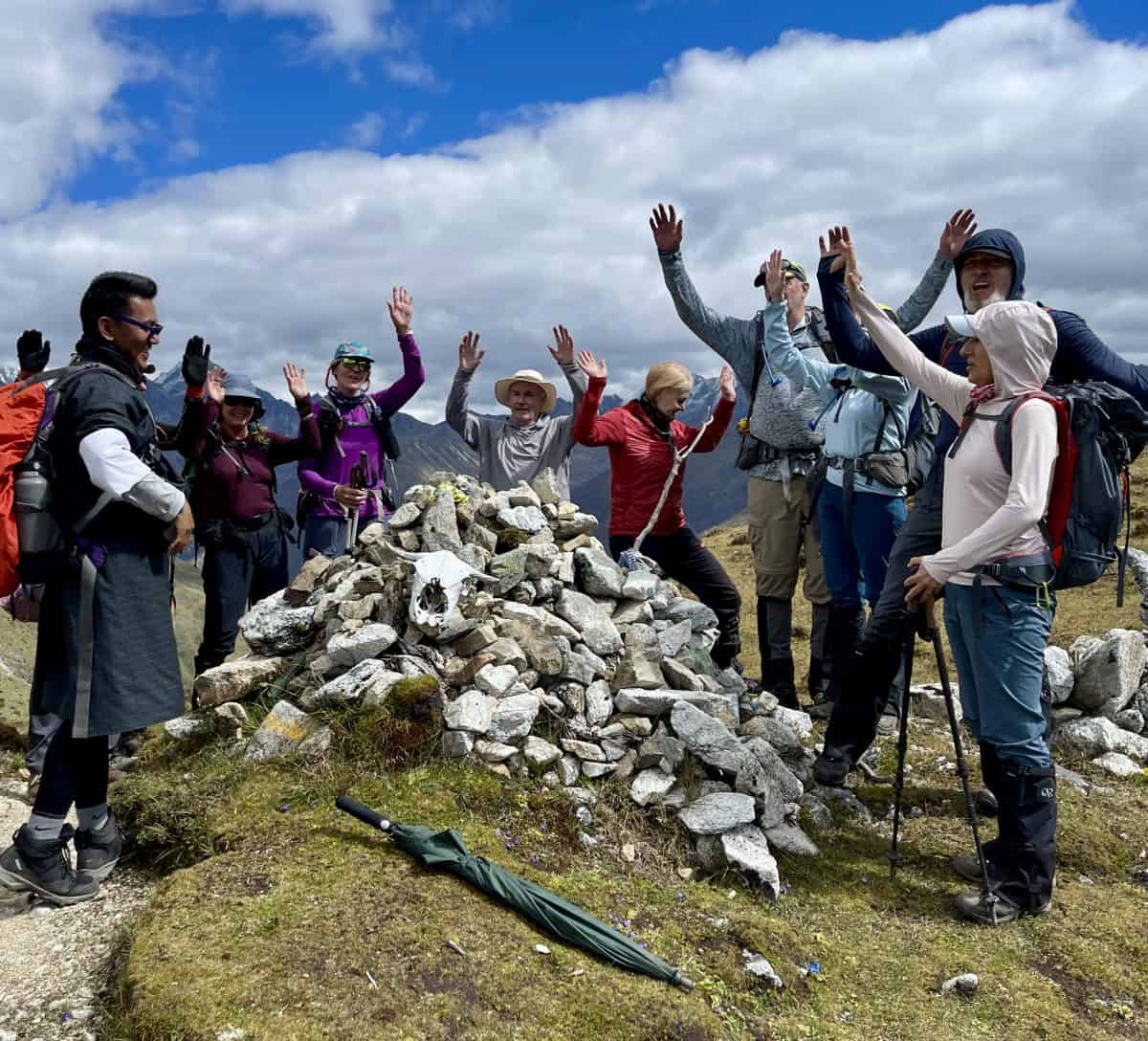 A group of hikers celebrates at a stone cairn on a mountain, raising their hands against a backdrop of dramatic clouds.