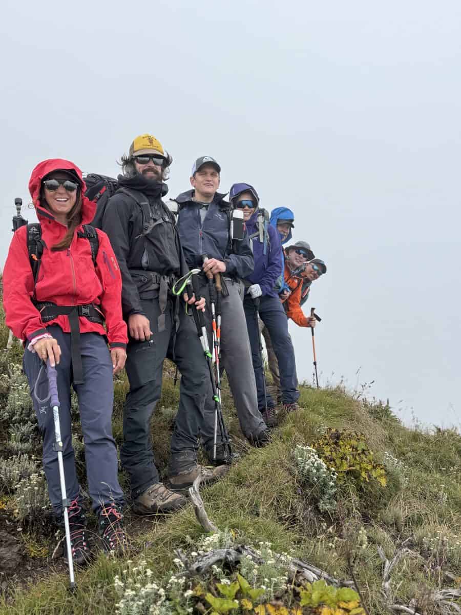 A group of six hikers stands on a hillside, dressed in outdoor gear, with foggy conditions surrounding them.