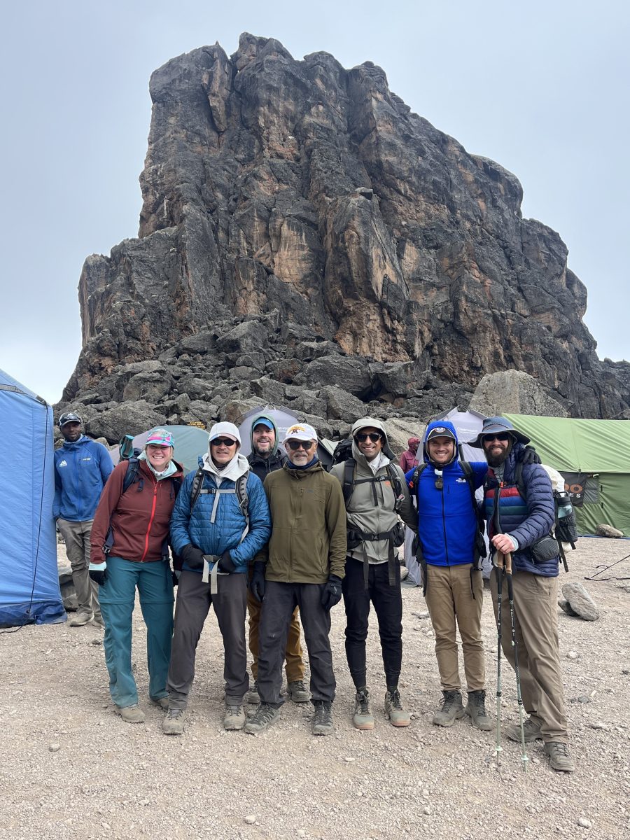 A group of seven hikers poses together in front of a large rock formation, with tents set up in a mountainous outdoor setting.