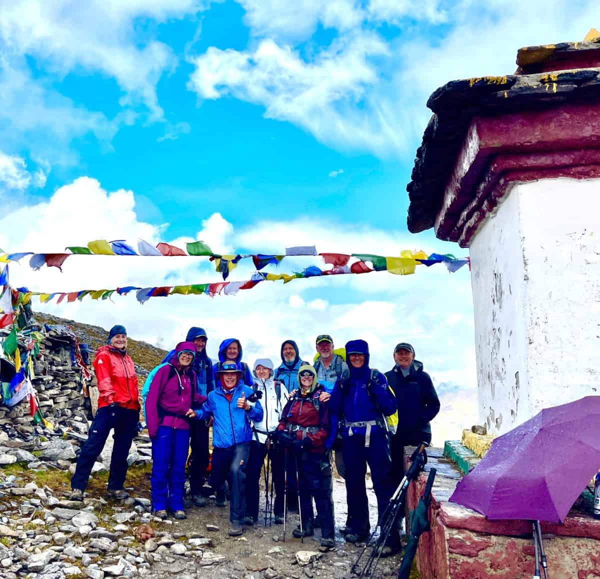 A group of hikers stands together near a stupa, surrounded by colorful prayer flags under a bright blue sky.