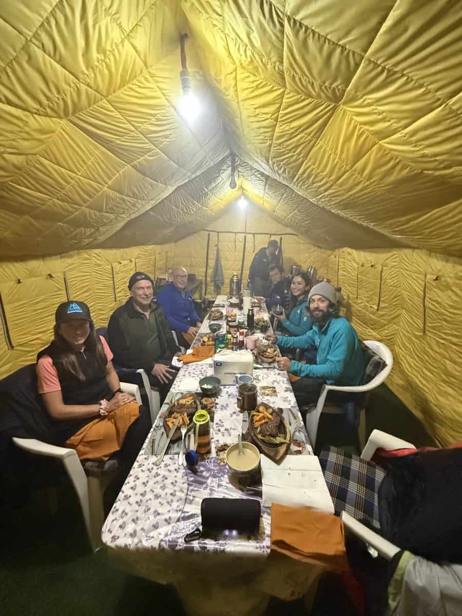 A group of people seated around a long table inside a large yellow tent, enjoying a meal together.