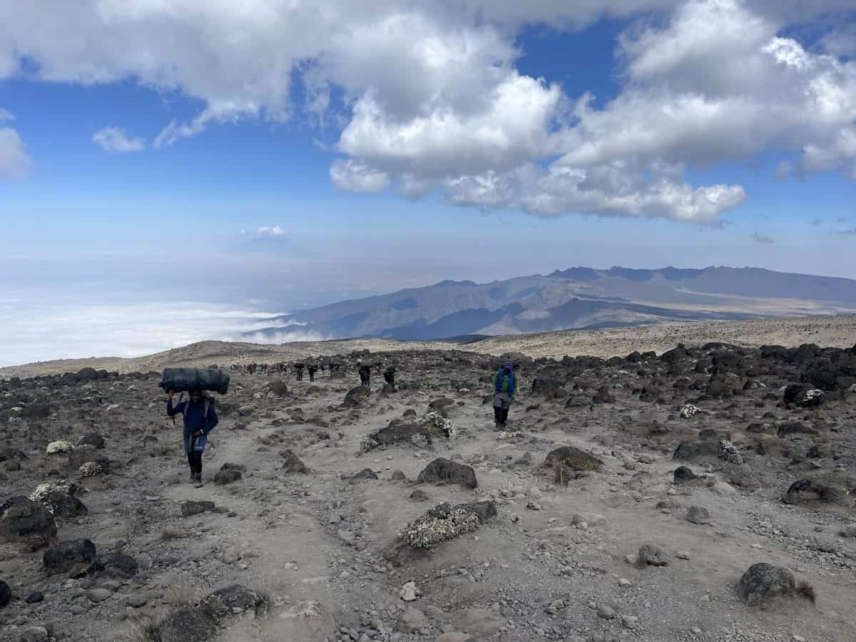 Hikers traverse a rocky landscape under a blue sky with white clouds, surrounded by mountains and a distant view of a vast valley.