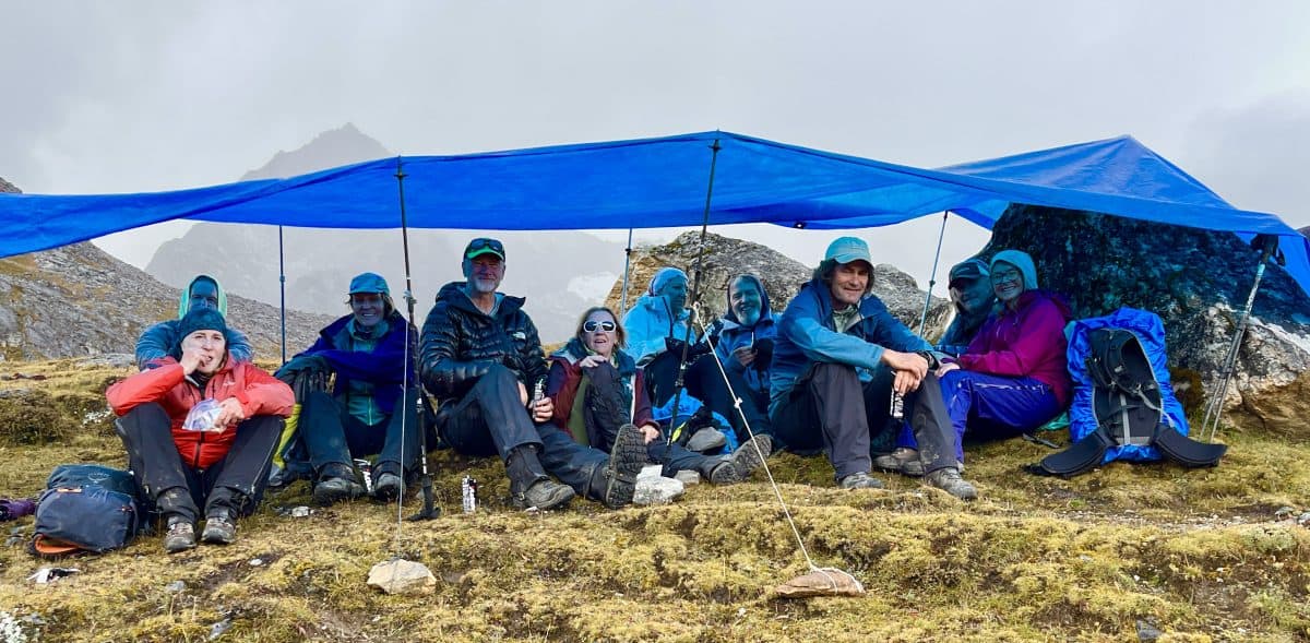 A group of people sheltering under a blue tarp in a mountainous area, wearing jackets and sitting on rocky ground.