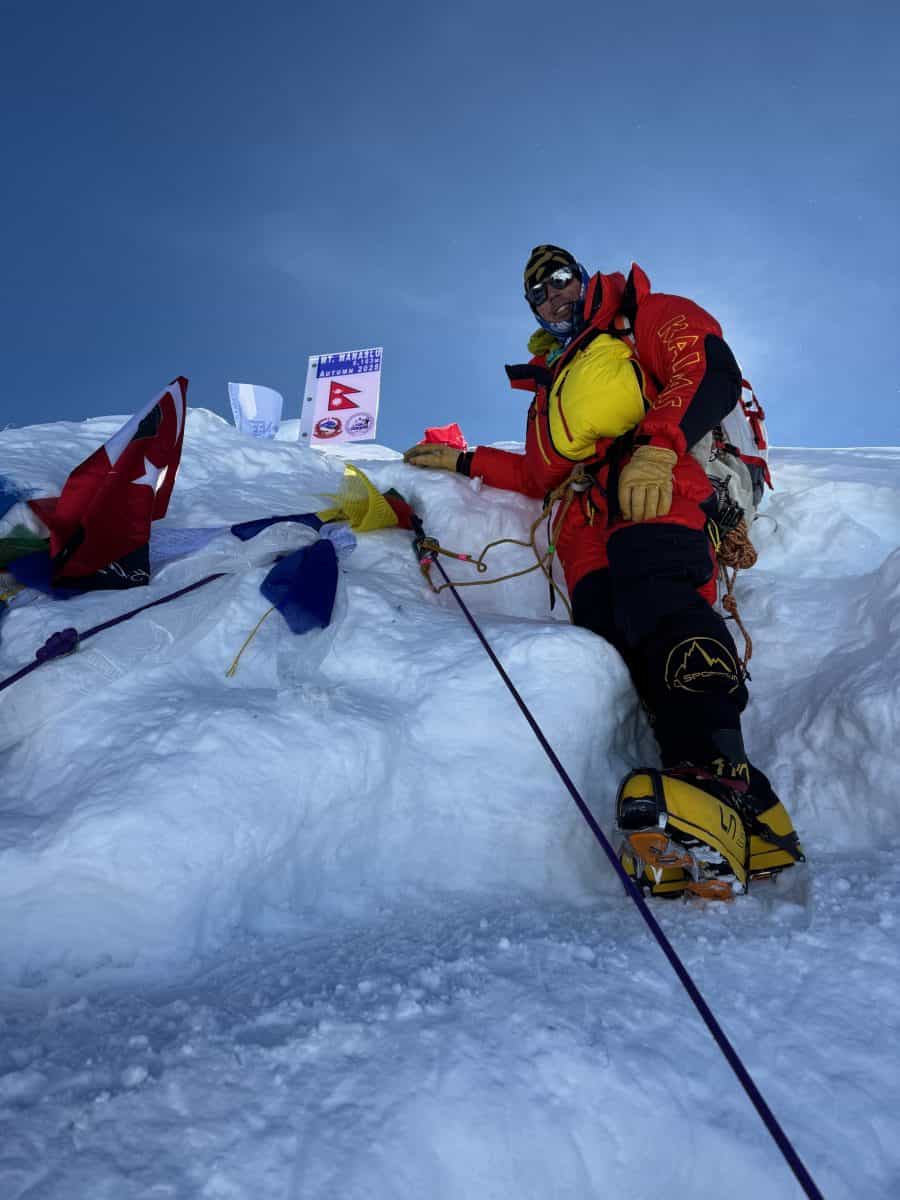 A climber stands on snow at a high-altitude peak, surrounded by flags and gear, showcasing determination and the thrill of mountaineering.