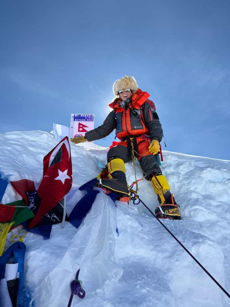 A climber stands on a snowy peak, surrounded by flags, celebrating at a mountaintop with a clear blue sky.