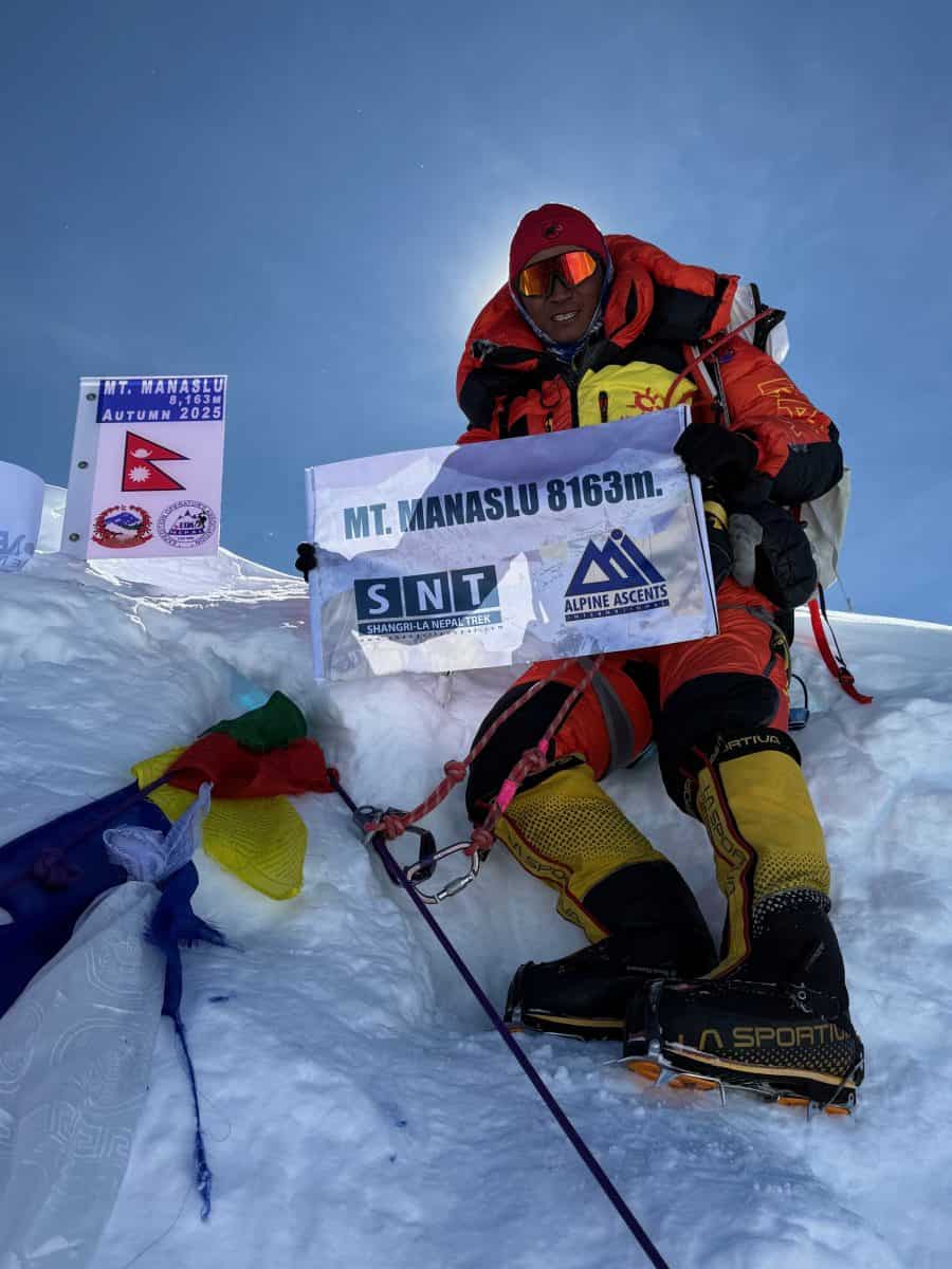 A climber sits on top of a snowy peak, holding a banner celebrating their ascent of Mount Manaslu, with colorful prayer flags in the foreground.