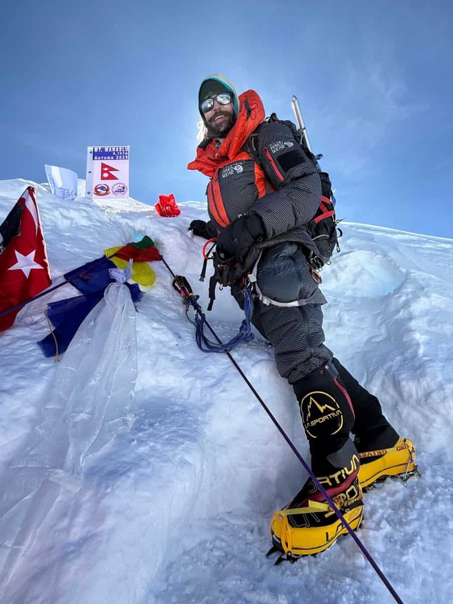 A climber stands triumphantly on a snowy peak, surrounded by flags, showcasing his achievement in high-altitude mountaineering.