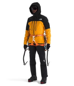 A person dressed in a bright orange and black climbing jacket with gear, including ice axes and a harness, preparing for an outdoor expedition.