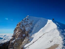 A snowy mountain peak under a clear blue sky, with two climbers trekking along the sharp ridge.
