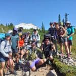 A group of hikers poses together in a scenic mountainous area, surrounded by greenery and towering trees, with a snow-capped peak in the background.