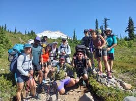 A group of hikers poses together in a scenic mountainous area, surrounded by greenery and towering trees, with a snow-capped peak in the background.