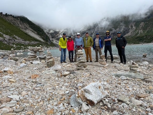 A group of seven hikers stands on rocky terrain, with a mountainous backdrop and a lake nearby, amidst overcast skies.