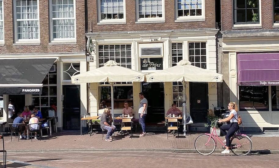 Fahrradfahrerin fährt vor sitzenden Menschen vor dem Café de Prins vorbei