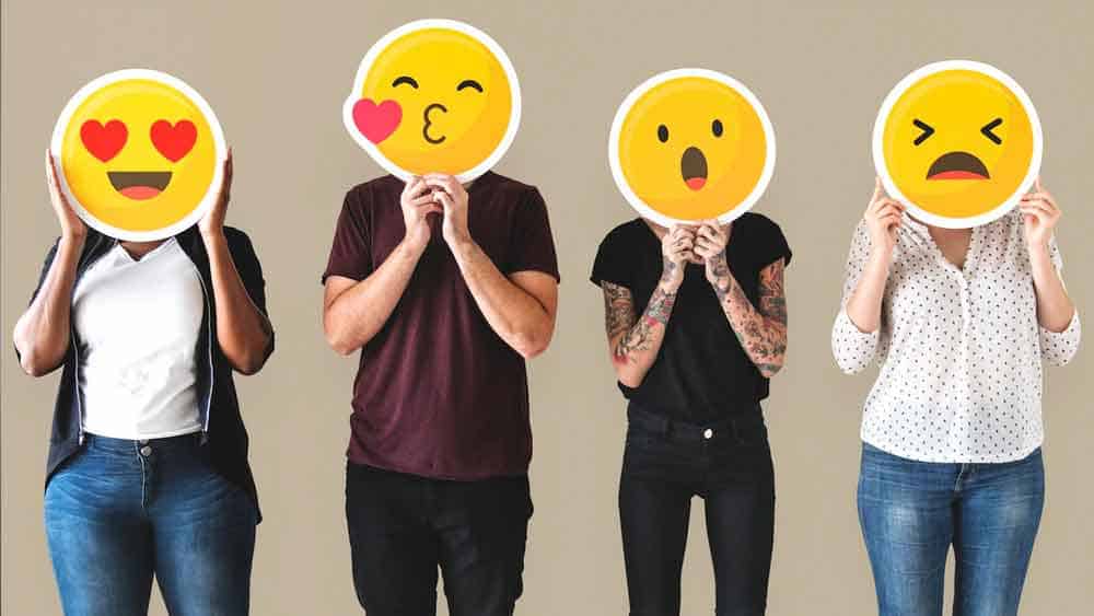 4 people holding emojis over their fact with different emotions