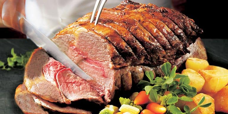 A person is carving a roasted meat joint with a knife and fork, accompanied by vegetables including potatoes and carrots.
