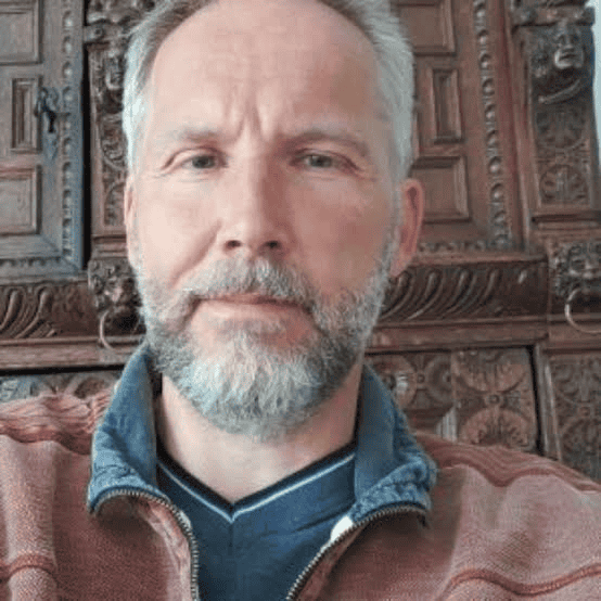 A middle-aged man with grey hair and a short beard is wearing a maroon jumper. He is sitting indoors in front of an ornately carved wooden background, looking at the camera with a neutral expression.