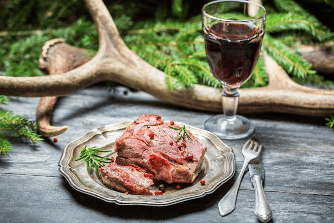 A plate with cooked venison garnished with rosemary and peppercorns sits beside a fork and knife, with a glass of red wine nearby. Antlers and pine branches are in the background on a rustic wooden table.