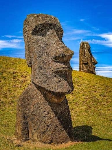 Two stone Moai statues stand on a grassy hillside under a blue sky.