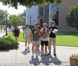 A group of young men outside on the sidewalk, with a woman walking up to them