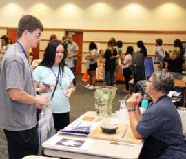 A male and female student by a table full of art projects, with professor in a T-shirt behind the table