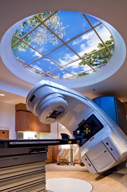 Large round window on ceiling of LINAC procedure room, featuring blue sky, clouds and flowering tree branches.