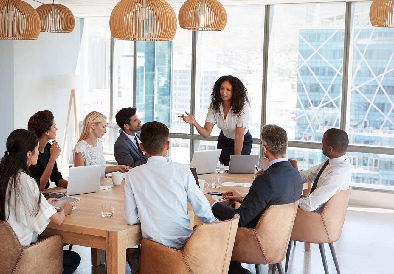 Group of people around a boardroom table with their attention on one person presenting